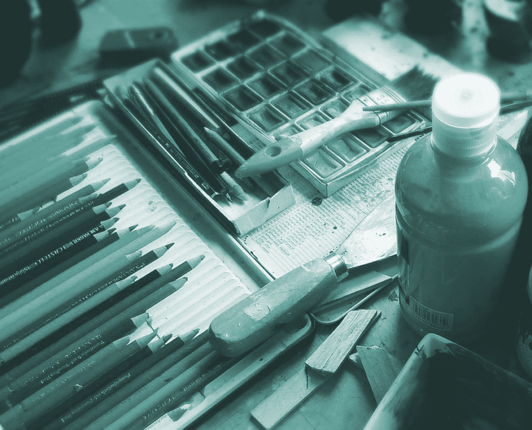 Man sieht eine Fotografie eines Arbeitsplatz mit Buntstiften, Farben, Pinsel und weiteres Kunstmaterial in einem Türkisstich.