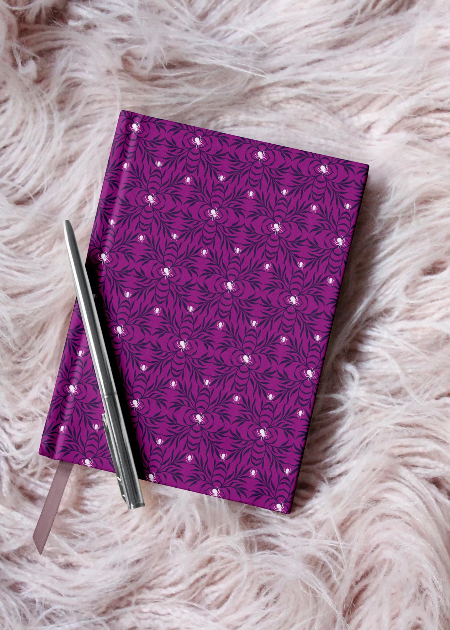 Notizbuchdesign von Vandyra Design mit floralen Spinnenmuster auf Kunstfell gebettet. Auf dem Notizbuch liegt ein silberner Kugelschreiber. Die Farben des Musters sind Lilatöne und weiße Spinnen. Der Hintergrund des Bildes ist das leicht rosafarbene Kunstfell.