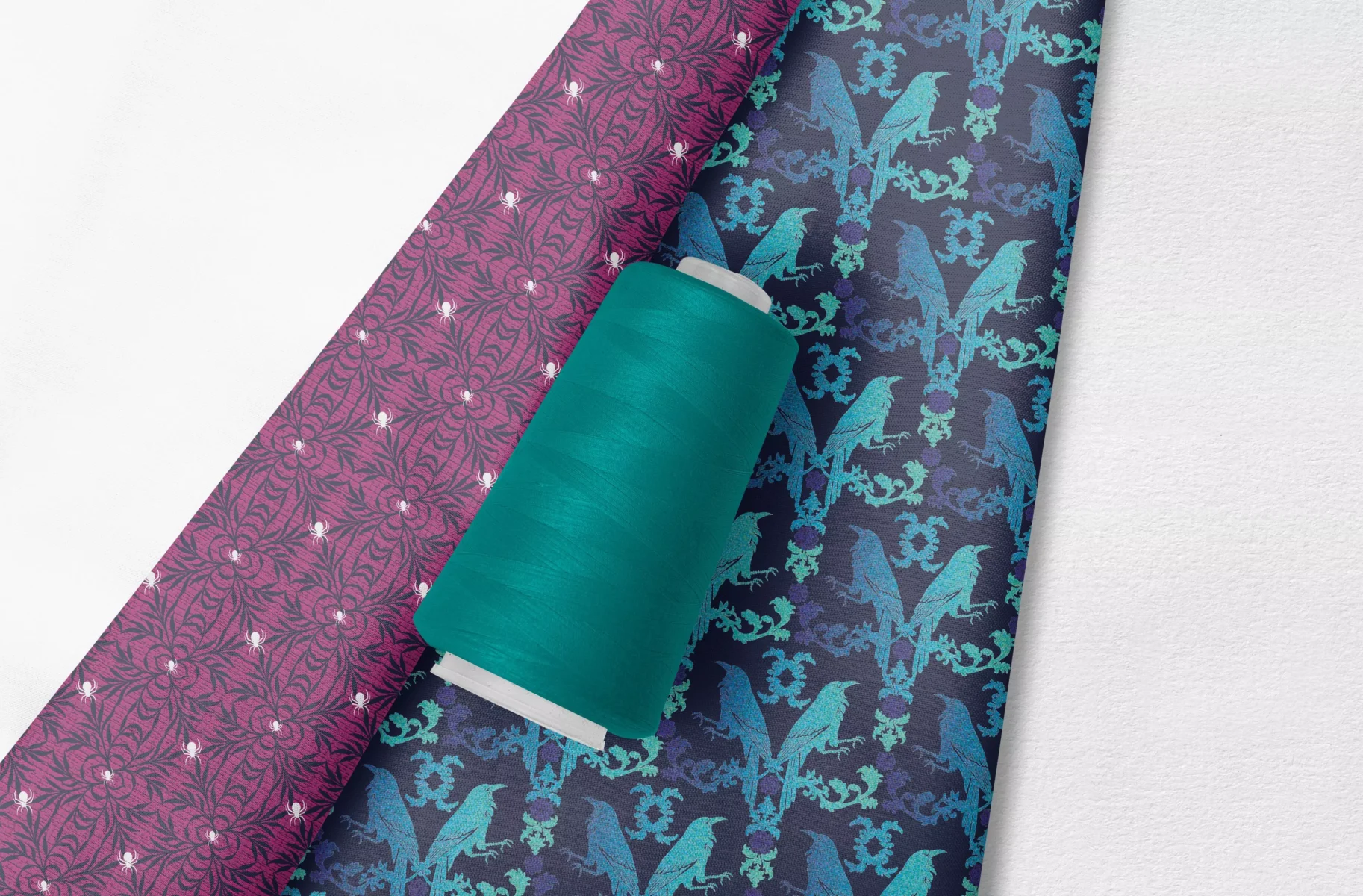 Hintergrundbild der Webseite von Vandyra Design. Zwei Stoffrollen. Ein florales Vogelmuster ist hellblau-lila mit Farbverlauf und ein florales Spinnenmuster rosa-lila mit weißen Spinnen und dazu ein türkisfarbenes Garn.