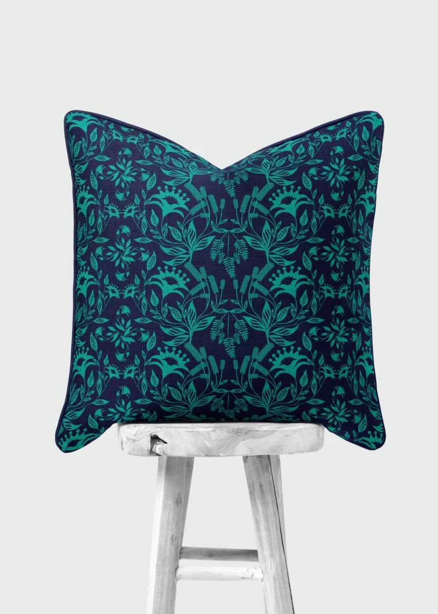 Stoffdesign von Vandyra Design. Ein Kissen mit türkisfarbenen Fantasiepflanzen und der Hintergrundstoff ein tiefes Nachtblau. Das Kissen steht auf einem weißen Holzstuhl mit rustikaler Holzmaserung. Der Bildhintergrund ist ein helles Grau.