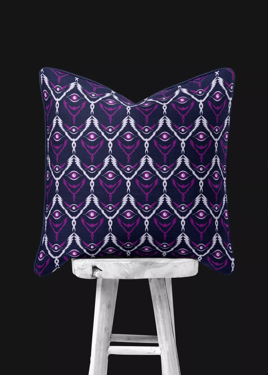 Stoffdesign von Vandyra Design. Ein Kissen mit mystischen fantasievollen flügelhaften Figuren die schweben. Farbspiel in Lilatönen und Lila-Grau. Hintergrund des Stoffes ein Mitternachtsblau. Das Kissen steht auf einem weißen Holzstuhl mit rustikaler Holzmaserung. Der Bildhintergrund ist ein Grau-Schwarz.