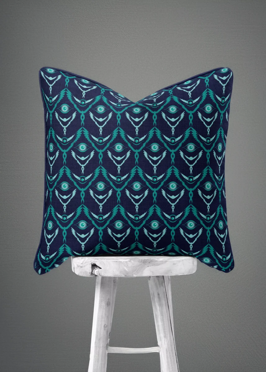 Stoffdesign von Vandyra Design. Ein Kissen mit mystischen fantasievollen flügelhaften Figuren die schweben. Farbspiel in verschiedenen Türkistönen von hell bis dunkel. Hintergrund des Stoffes ein Mitternachtsblau. Das Kissen steht auf einem weißen Holzstuhl mit rustikaler Holzmaserung. Der Bildhintergrund ist ein Grau-Schwarz.