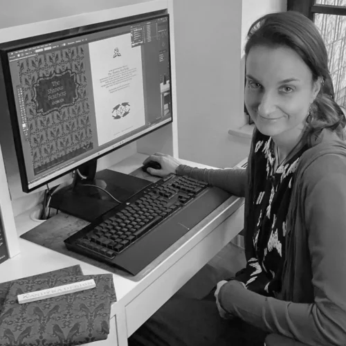 Portrait der Designerin Vandyra Design. Sie sitzt an ihrem Arbeitsplatz, vor ihr der eingeschaltete PC und dieser Bildschirm zeigt eine ihrer Kollektionen. Die Aufnahme ist in Schwarz-Weiß.